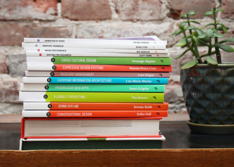 Stack of books next to a green plant, in front of a brick wall.
