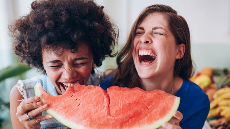 Two women laugh and bite into a large piece of watermelon.