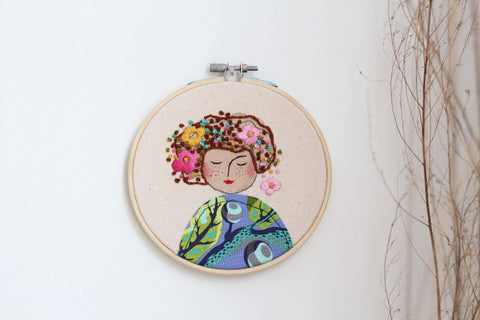 Embroidery of an auburn-haired woman with periwinkle fabric hanging on a wall