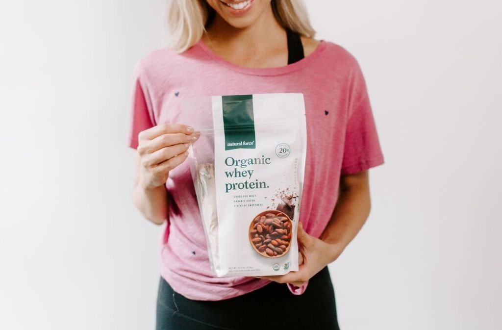 fit woman smiling while holding a bag of natural force organic whey protein