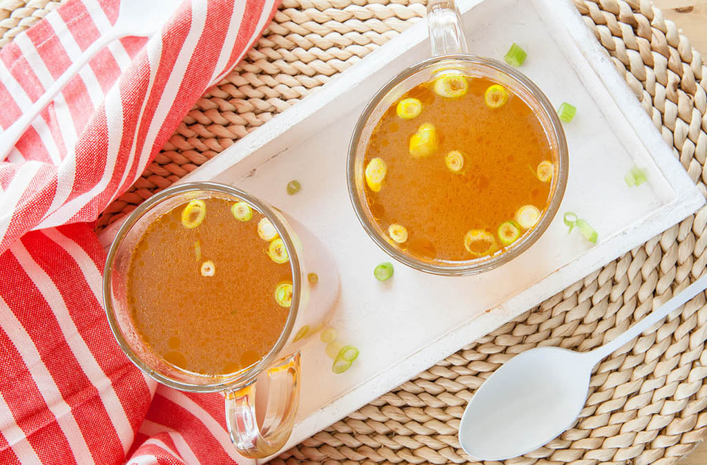 two mugs of natural forces fast bone broth recipe on a white rectangular tray next to a red and white striped dish towel