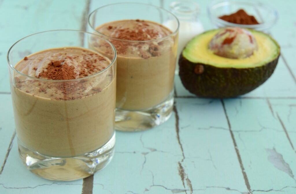 two glasses of chocolate avocado smoothie with almond butter and protein beside half an avocado on a teal wooden surface