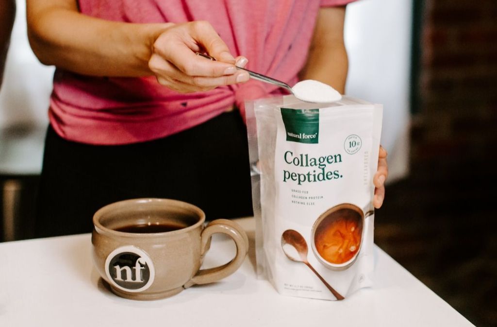spoon scooping collagen peptides out of a natural force grass fed collagen peptides bag beside a mug of clean coffee