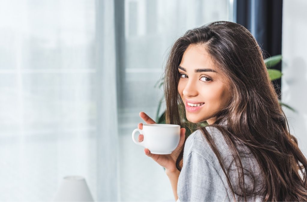 woman with thick shiny hair looking over her shoulder and smiling while holding a cup of coffee