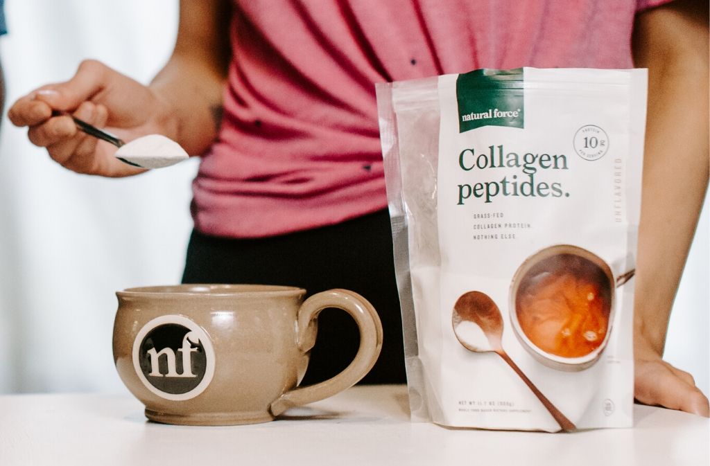 hand holding a spoonful of collagen peptides over a handcrafted natural force latte mug beside a bag of natural force collagen peptides