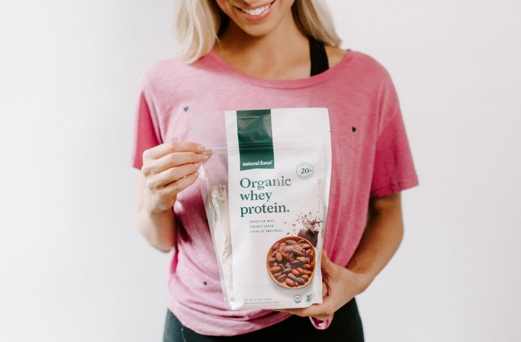 athletic woman in a pink shirt smiling while holding a bag of natural force organic whey protein