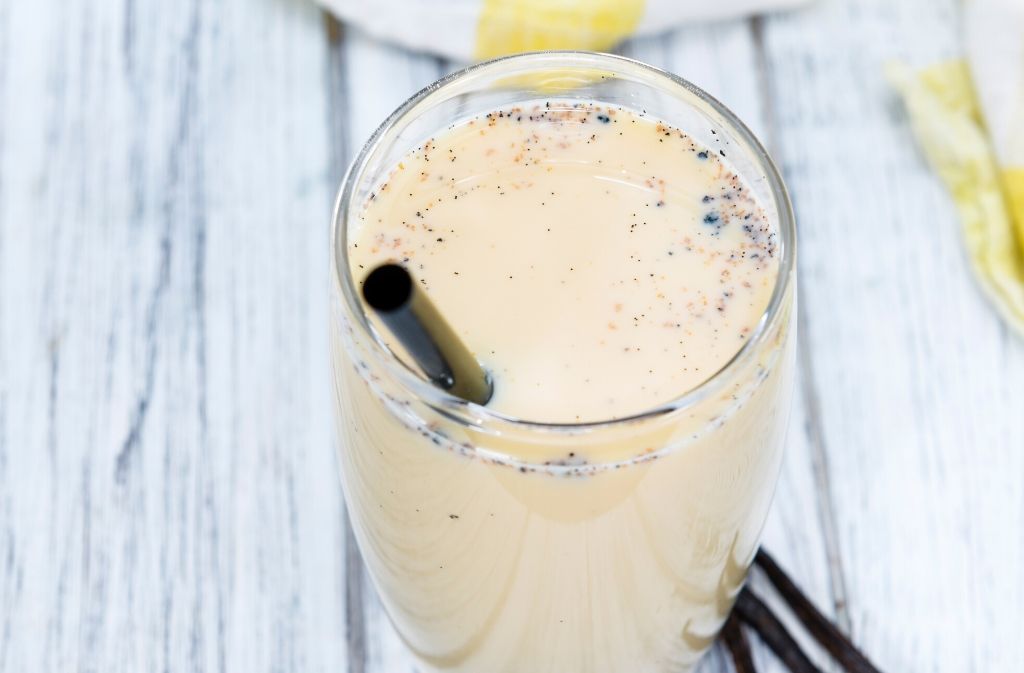 a glass of lemon cake keto shake on a white washed wood surface in front of a yellow and white striped hand towel