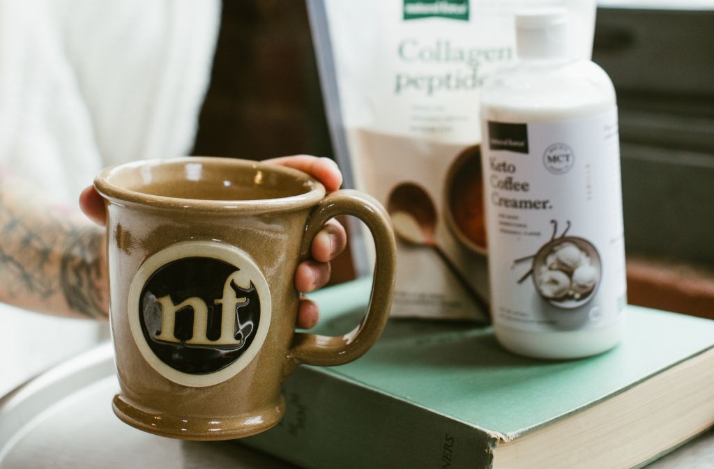 hand holding out a handmade natural force mug next to natural force collagen peptides and keto coffee creamer