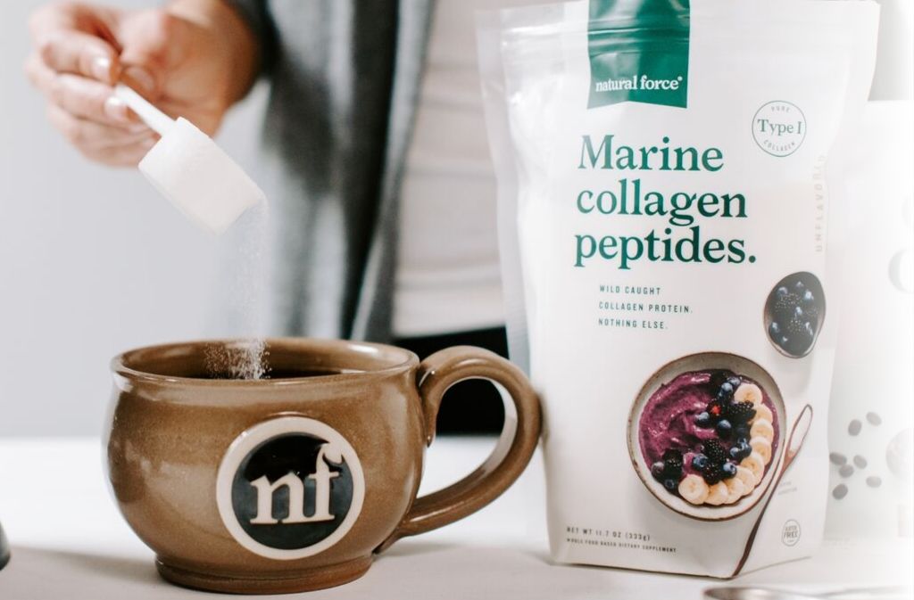 hand pouring marine collagen peptides into a handcrafted natural force mug of keto chai tea latte beside a bag of natural force marine collagen peptides