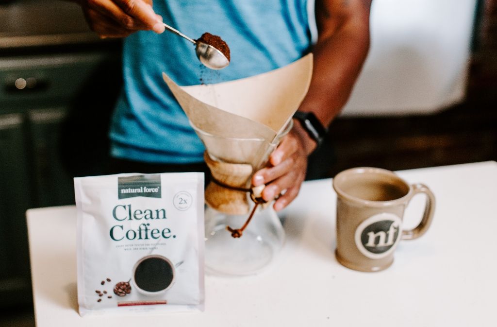 man pouring a tablespoon of ground coffee into a chemex beside a bag of natural force clean coffee and a handmade natural force mug