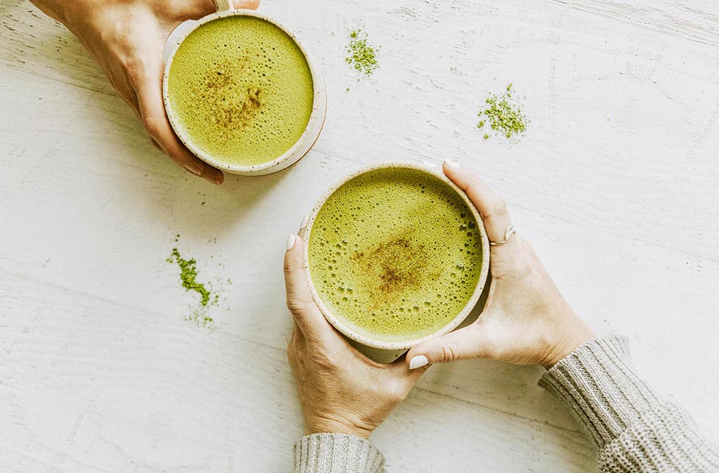 hands holding two mugs of keto matcha latte on a white wood surface