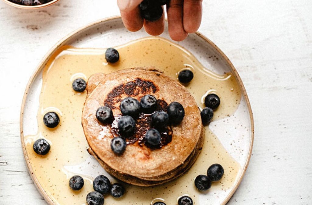 hand dropping blueberries on a plat of paleo protein pancakes