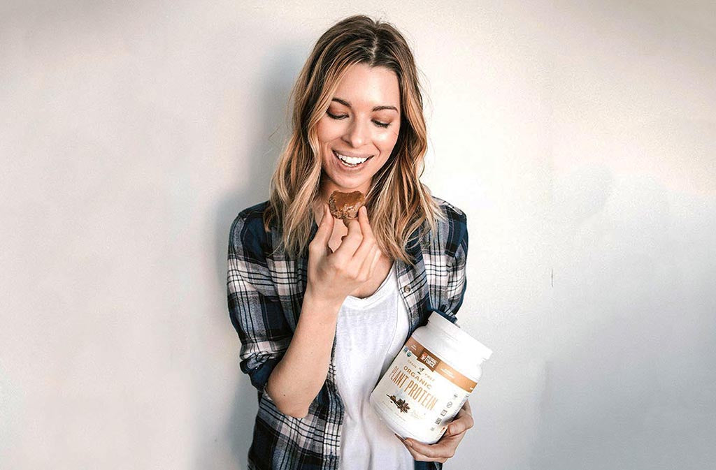 blond woman smiling about to take a bite of keto cookie holding a container of organic vegan plant protein