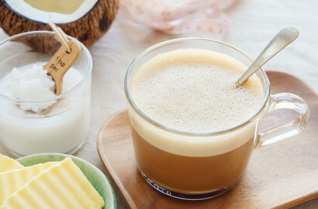 a clear glass mug of frothy keto coffee on a wooden plate surrounded by grass fed butter mct oil and coconut