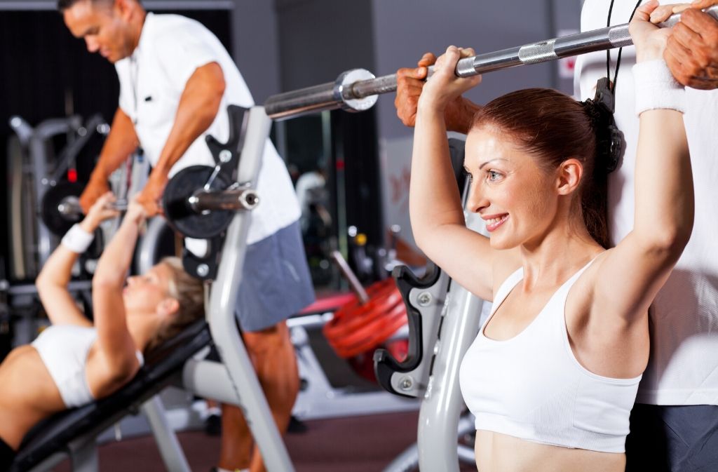 athletic woman in a white sports bra smiling while doing a seated shoulder press while a man stands behind her lightly supporting the weight of the bar