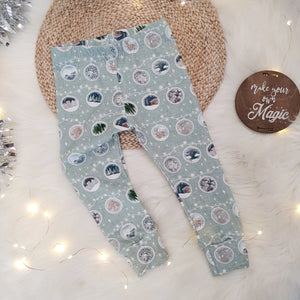 Christmas bauble printed leggings by bayridgecaskandkeg. These baby and child leggings feature a light green background with garlands of baubles each depicting a different wintery scene. The leggings are pictured against a white, fluffy rug with wicker placemat, silver tinsel and fairy lights to accent.