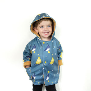 Little boy wearing a bayridgecaskandkeg pigeon puddles splasher jacket. The jacket is blue, with pigeons wearing yellow rain coats, hats and wellies. The fabric also features umbrellas and raindrops. 