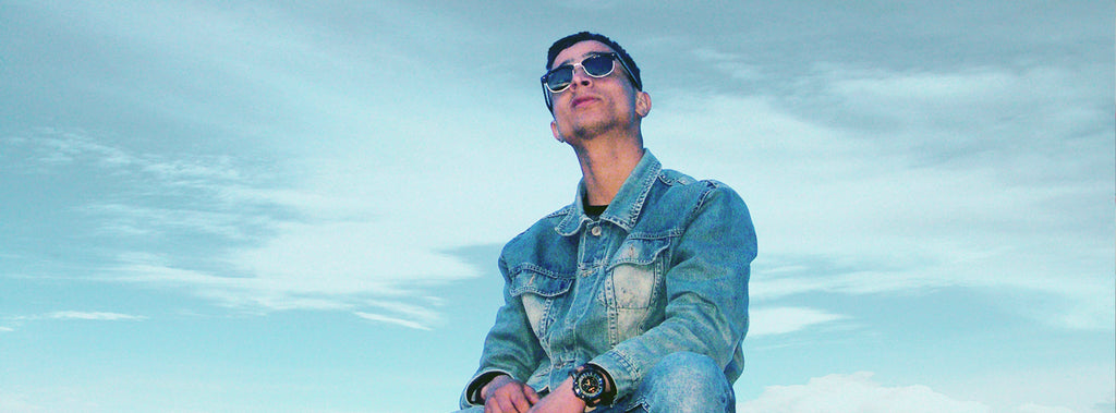 Man wearing sunglasses and denim jacket looking up at the sky.