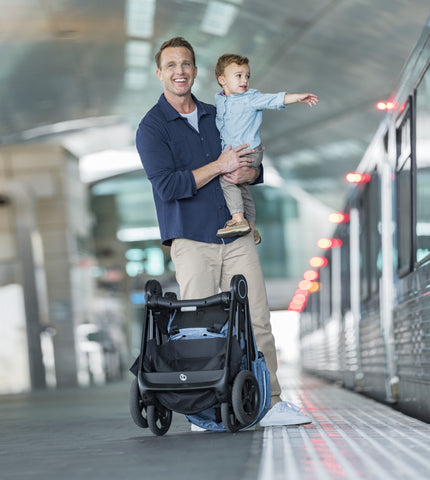 Man holding child while standing behind folded Stokke Beat on a train platform