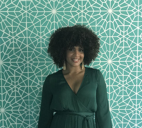 A woman in a dark green dress standing in front of a decorative wall with a teal and white pattern, paint colours to brighten up your space.