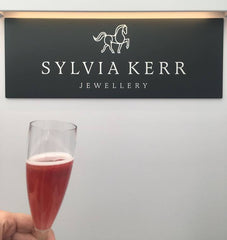 Champagne flute of cocktail infront of Sylvia kerr Jewellery Sign