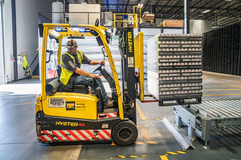 man reversing yellow forklift truck carrying a pallet of goods in a warehouse 