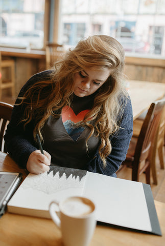 Shelby Campbell sitting in Leavenworth coffee shop wearing a grey graphic t-shirt with a heart and mountain scene illustration. PNW Style. Pacific Northwest. Drawing. Sipping Coffee. Cafe.  Leavenworth, Washington. Mountains. Mountain Style.