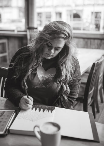 Shelby Campbell sitting in Leavenworth coffee shop wearing a grey graphic t-shirt with a heart and mountain scene illustration. PNW Style. Pacific Northwest. Drawing. Sipping Coffee. Cafe.  Leavenworth, Washington. Mountains. Mountain Style.