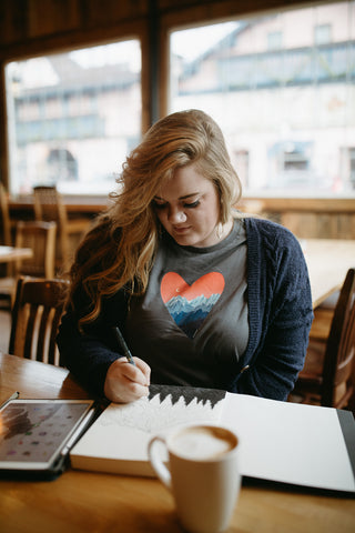 Shelby Campbell sitting in Leavenworth coffee shop wearing a grey graphic t-shirt with a heart and mountain scene illustration. PNW Style. Pacific Northwest. Drawing. Sipping Coffee. Cafe.  Leavenworth, Washington. Mountains. Mountain Style.