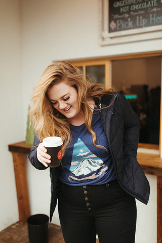 Women wearing a blue graphic t-shirt with a triangle shaped mountain illustration on the front. Black denim jeans. High Waisted. Booties. Coffee. Outdoor Coffee Shop in Leavenworth, Washington. PNW Style. Pacific Northwest.