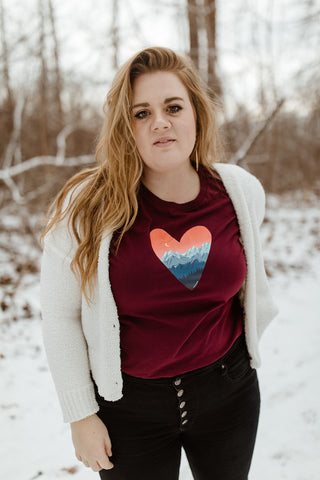 Mountain Love Graphic T-shirt. Tee. Mountain and Heart Illustration. Woman wearing white sweater, black pants, and brown booties in a snowy forested area in Leavenworth, Washington. PNW Style. Pacific Northwest. Casual Style. Mountain Style. Mountains.