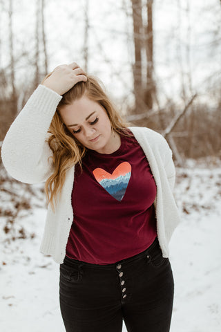 Mountain Love Graphic T-shirt. Tee. Mountain and Heart Illustration. Woman wearing white sweater, black pants, and brown booties in a snowy forested area in Leavenworth, Washington. PNW Style. Pacific Northwest. Casual Style. Mountain Style. Mountains.