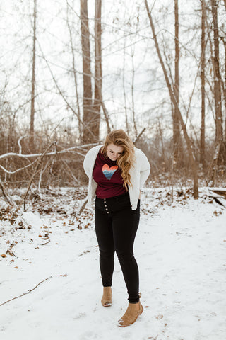 Mountain Love Graphic T-shirt. Tee. Mountain and Heart Illustration. Woman wearing white sweater, black pants, and brown booties in a snowy forested area in Leavenworth, Washington. PNW Style. Pacific Northwest. Casual Style. Mountain Style. Mountains.