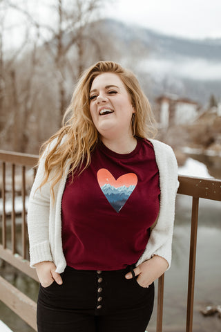 Mountain Love Graphic T-shirt. Tee. Mountain and Heart Illustration. Woman wearing white sweater, black pants, and brown booties in a snowy forested area in Leavenworth, Washington. PNW Style. Pacific Northwest. Casual Style. Mountain Style. Mountains.