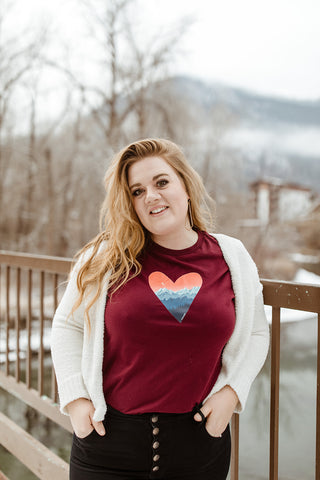 Mountain Love Graphic T-shirt. Tee. Mountain and Heart Illustration. Woman wearing white sweater, black pants, and brown booties in a snowy forested area in Leavenworth, Washington. PNW Style. Pacific Northwest. Casual Style. Mountain Style. Mountains.