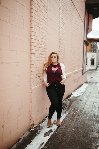 Mountain Love Graphic T-shirt. Tee. Mountain and Heart Illustration. Woman wearing white sweater, black pants, and brown booties in a snowy forested area in Leavenworth, Washington. PNW Style. Pacific Northwest. Casual Style. Mountain Style. Mountains.