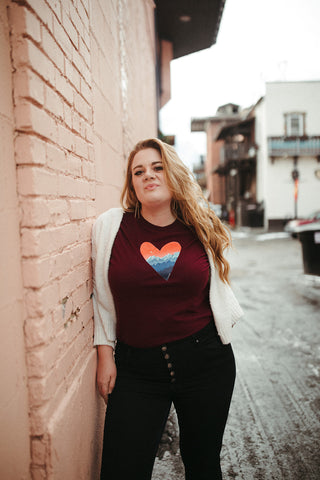 Mountain Love Graphic T-shirt. Tee. Mountain and Heart Illustration. Woman wearing white sweater, black pants, and brown booties in a snowy forested area in Leavenworth, Washington. PNW Style. Pacific Northwest. Casual Style. Mountain Style. Mountains.