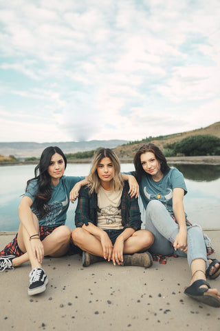 Group of women sitting in front of a small lake/pond. Wearing graphic t-shirts with a steer skull design on the front. PNW style. Wenatchee, WA. Pacific Northwest. Graphic Tee.