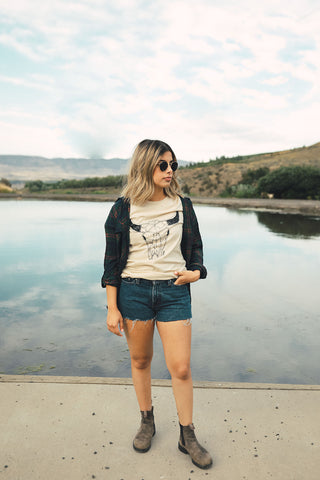 Woman standing in front of small lake/pond in Wenatchee, WA. Wearing a graphic t-shirt with a steer skull design on the front. Jeans Shorts. Round Sunglasses. Summer Time.PNW Style. Pacific Northwest.