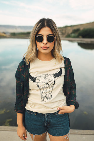 Woman standing in front of small lake/pond in Wenatchee, WA. Wearing a graphic t-shirt with a steer skull design on the front. Jeans Shorts. Round Sunglasses. Summer Time.PNW Style. Pacific Northwest.