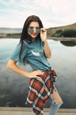 Woman standing in front of small lake/pond in Wenatchee, WA. Wearing a graphic t-shirt with a steer skull design on the front. Jeans. Flannel Shirt. Round Sunglasses. Summer Time.PNW Style. Pacific Northwest.