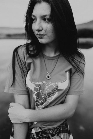 Woman standing in front of small lake/pond in Wenatchee, WA. Wearing a graphic t-shirt with a steer skull design on the front. Jeans. Flannel Shirt. Round Sunglasses. Summer Time.PNW Style. Pacific Northwest.