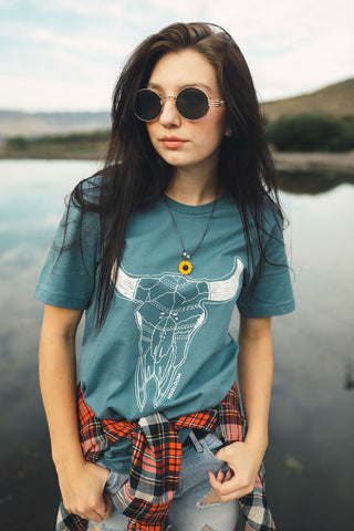 Woman standing in front of small lake/pond in Wenatchee, WA. Wearing a graphic t-shirt with a steer skull design on the front. Jeans. Flannel Shirt. Round Sunglasses. Summer Time.PNW Style. Pacific Northwest.