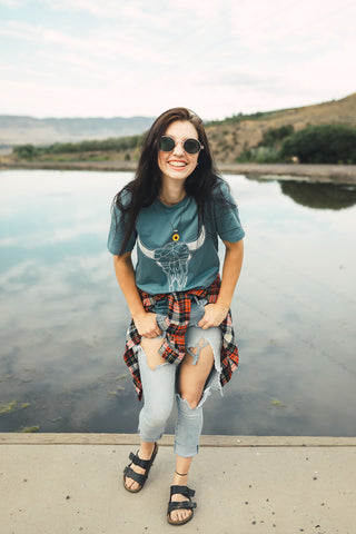 Woman standing in front of small lake/pond in Wenatchee, WA. Wearing a graphic t-shirt with a steer skull design on the front. Jeans. Flannel Shirt. Round Sunglasses. Summer Time.PNW Style. Pacific Northwest.