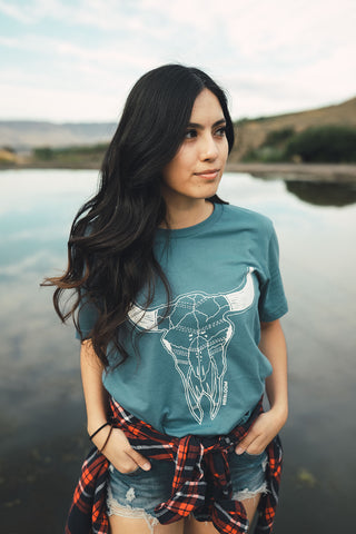 Woman standing in front of small lake/pond in Wenatchee, WA. Wearing a graphic t-shirt with a steer skull design on the front. Jeans. Flannel Shirt. Round Sunglasses. Summer Time.PNW Style. Pacific Northwest.