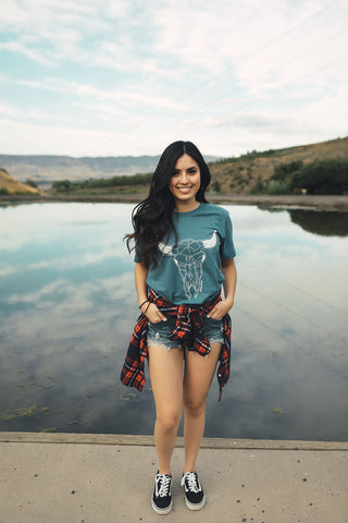 Woman standing in front of small lake/pond in Wenatchee, WA. Wearing a graphic t-shirt with a steer skull design on the front. Jeans. Flannel Shirt. Summer Time.PNW Style. Pacific Northwest.