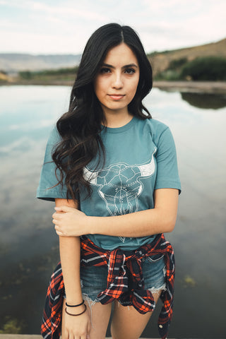 Woman standing in front of small lake/pond in Wenatchee, WA. Wearing a graphic t-shirt with a steer skull design on the front. Jeans. Flannel Shirt. Summer Time.PNW Style. Pacific Northwest.