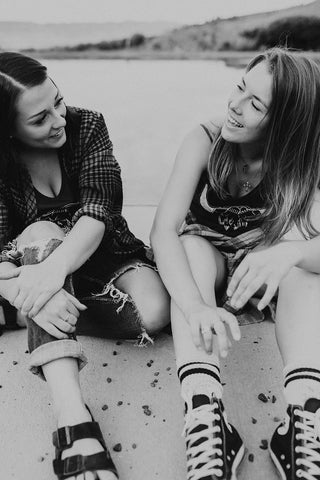 Two women sitting in front of small lake/pond wearing graphic tank tops with a steer skull design on the front. Also wearing flannel shirts, jeans, and converse and birkenstock sandals. PNW Style. Pacific Northwest. Wenatchee, Washington. Mountain Style. Golden Hour.