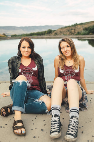 Two women sitting in front of small lake/pond wearing graphic tank tops with a steer skull design on the front. Also wearing flannel shirts, jeans, and converse and birkenstock sandals. PNW Style. Pacific Northwest. Wenatchee, Washington. Mountain Style. Golden Hour.