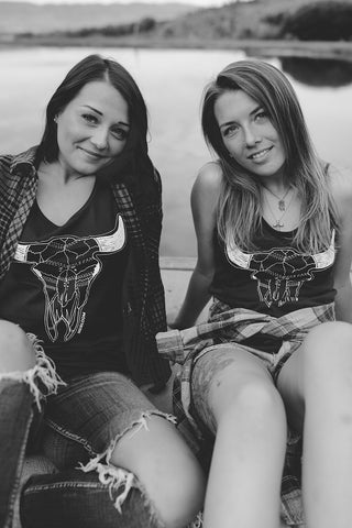 Two women sitting in front of small lake/pond wearing graphic tank tops with a steer skull design on the front. Also wearing flannel shirts, jeans, and converse and birkenstock sandals. PNW Style. Pacific Northwest. Wenatchee, Washington. Mountain Style. Golden Hour.